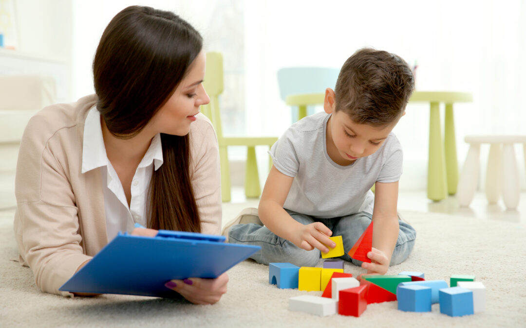 Young child psychologist working with little boy, playing on the floor with colorful blocks
